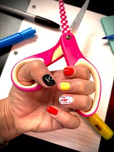 Colorful nails holding pink scissors, stationery background.